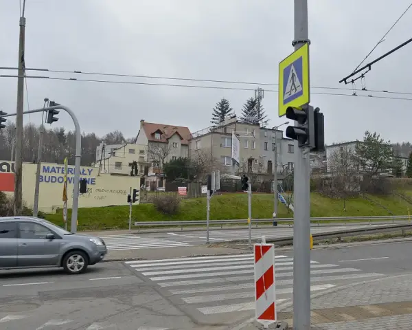 Szare auto zbliża się do przejścia dla pieszych, gdzie światła kolizyjne regulują ruch. W tle budynki i drzewa.