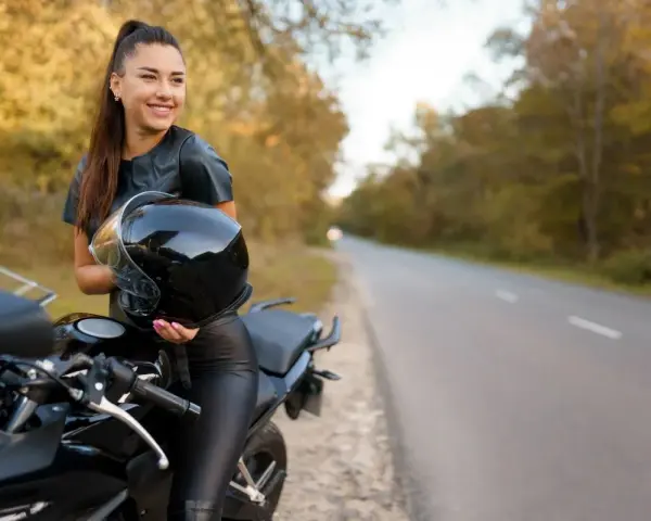 Uśmiechnięta kobieta w skórzanym stroju trzyma kask obok swojego motocykla. Idealne motory dla kobiet na jesienną przejażdżkę.