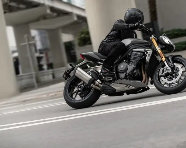 Motocyklista w kasku i skórzanej kurtce pochyla się na swoim motocyklu Triumph Speed Triple, pokonując zakręt na miejskiej drodze.