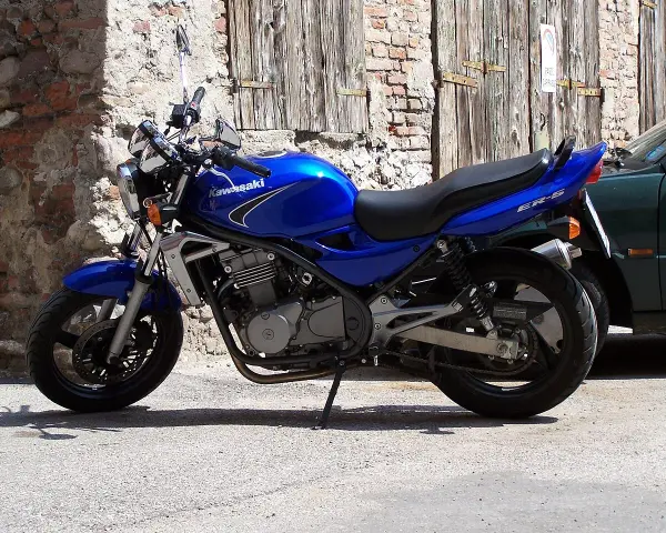 Błękitny motocykl Kawasaki ER-5 zaparkowany przy kamiennym murze i drewnianej bramie.