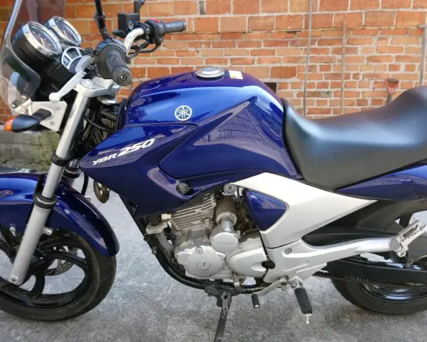 Błękitny motocykl Yamaha YBR 250 z chromowanymi elementami, zaparkowany na tle ceglanej ściany.