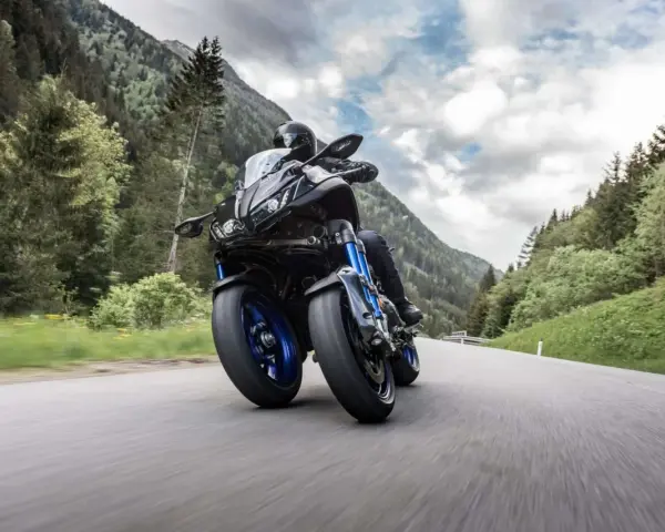 Motocyklista na Yamaha Niken pokonuje zakręt na górskiej drodze, otoczony zielonymi lasami i skalistymi szczytami.
