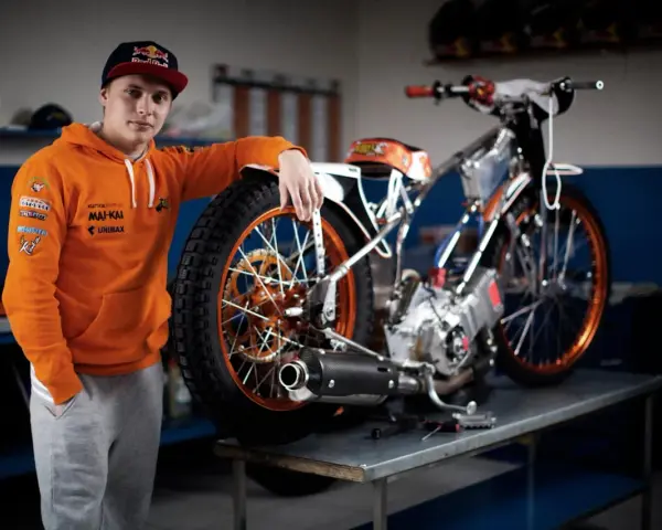 Młody żużlowiec w pomarańczowej bluzie opiera się o swój motocykl żużlowy, gotowy do kolejnego wyścigu.