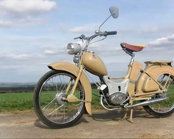 Beżowy motorower z NRD, z charakterystycznym siodełkiem i błotnikami, stoi na tle zielonych pól i błękitnego nieba.