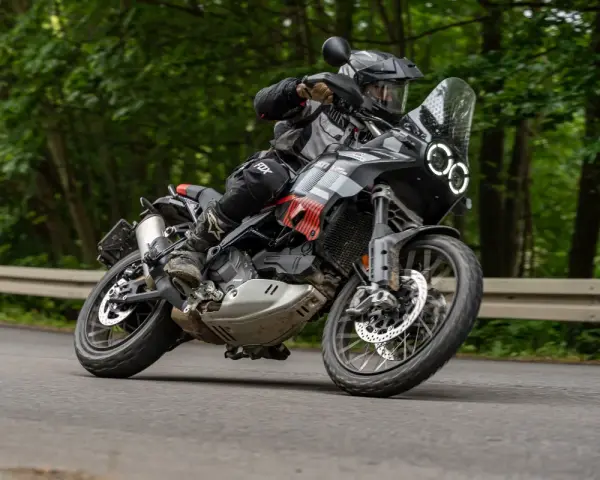 Motocyklista w kasku i stroju terenowym pochyla się na zakręcie na drodze leśnej, prowadząc czarny motocykl Ducati DesertX.