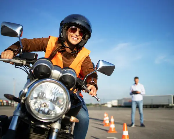 Uśmiechnięta kursantka w kamizelce odblaskowej i kasku na motocyklu, przygotowuje się do egzaminu na prawo jazdy A2.