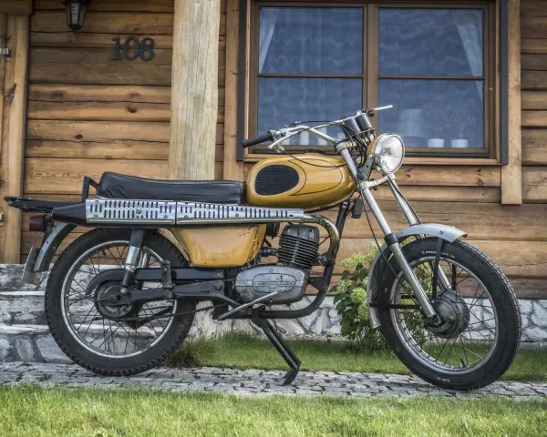 Żółty motorower WSK 175, z widocznymi danymi technicznymi na silniku, stoi przed drewnianym domem.