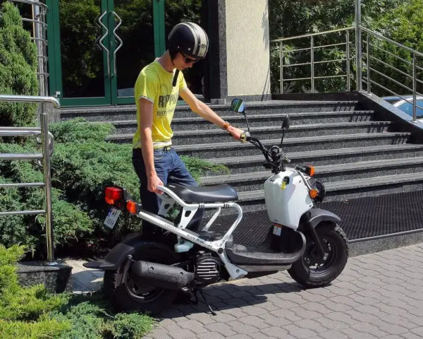 Młody mężczyzna w kasku i żółtej koszulce przy białym skuterze. To jego stara karta motorowerowa, gotowa do jazdy.