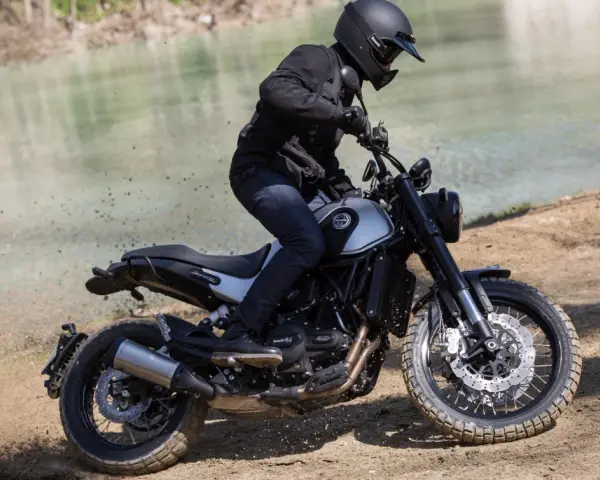 Motocyklista w kasku i czarnej kurtce jedzie motocyklem Benelli Leoncino 500 Trail po piaszczystym brzegu rzeki, wzbijając tumany kurzu.