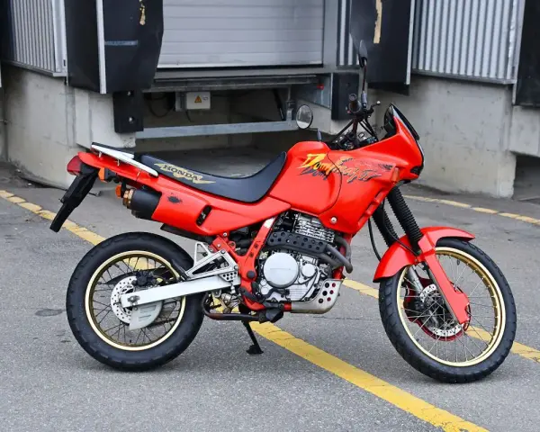 Czerwony motocykl Honda Dominator z lat 90. stoi na asfalcie. Ma złote felgi i czarne siedzenie.
