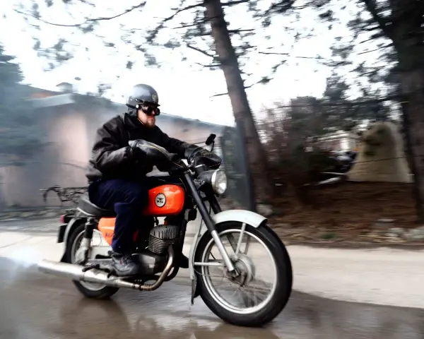Mężczyzna w kasku i goglach jedzie na pomarańczowym motocyklu WSK. Widać dym z rury wydechowej.