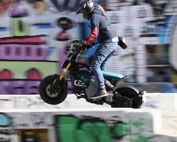 Młody człowiek na elektrycznym motocyklu BMW CE02 skacze nad przeszkodą na tle kolorowego graffiti.