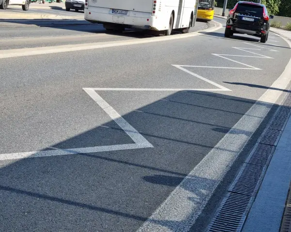 Na drodze zygzakowata linia, znak p-17, zapowiada zbliżanie się do przejścia dla pieszych. W tle autobus i samochód.