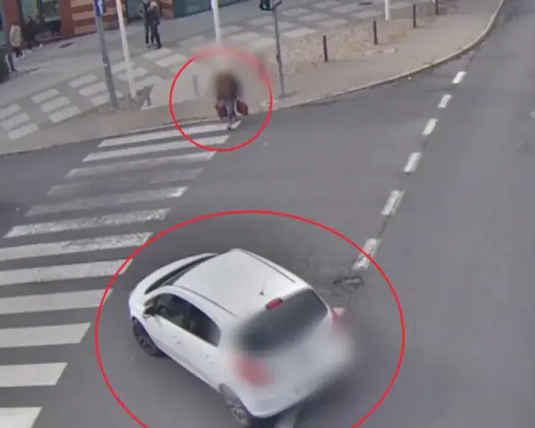 Biały samochód wymusza pierwszeństwo na pasach, omal nie potrącając pieszego z walizką. Inny samochód zatrzymał się przed przejściem.