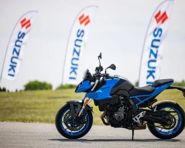 Niebieski motocykl Suzuki GSX-S z niebieskimi felgami stoi na asfalcie. W tle powiewają białe flagi z logo Suzuki.