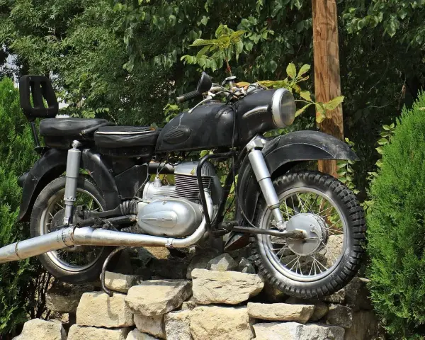 Czarny motocykl z matowym malowaniem baku, stojący na kamiennym murku wśród zieleni.