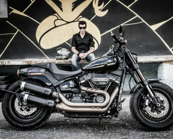 Mężczyzna w czarnej koszuli i jeansach siedzi obok lśniącego, czarnego motocykla Harley-Davidson Fat Bob.