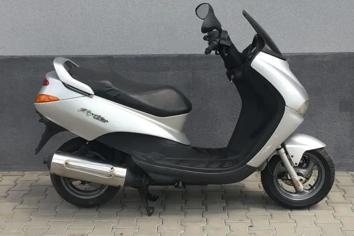 Srebrny skuter Peugeot Elyseo 125 z czarnym siedzeniem i przyciemnianą szybą, zaparkowany na chodniku.