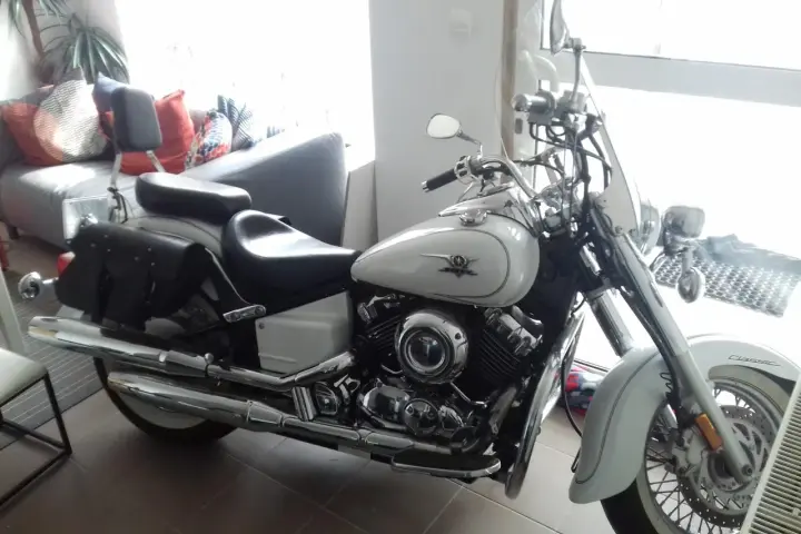 Biały motocykl Yamaha Drag Star 650, z sakwami i szybą. Wady i zalety tego klasyka są dyskutowane w tle, gdzie widać kanapę.