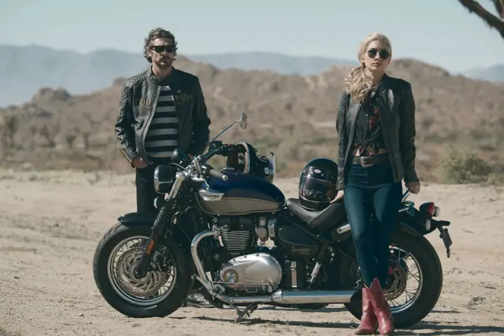 Para w skórzanych kurtkach pozuje przy motocyklu Triumph Speedmaster na pustynnym tle.