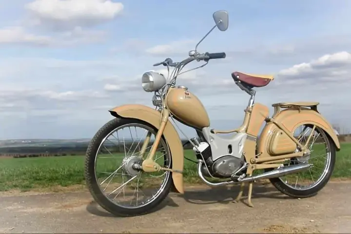Beżowy motorower z NRD, z charakterystycznym siodełkiem i błotnikami, stoi na tle zielonych pól i błękitnego nieba.