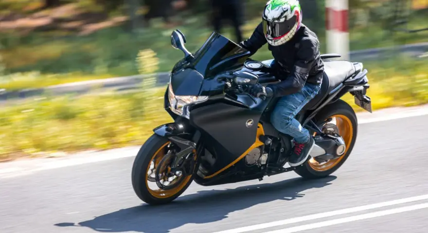 Motocyklista w kasku z uśmiechniętą twarzą pędzi na czarnym motocyklu Honda VFR 1200 po drodze.