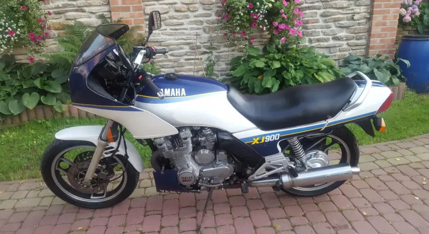 Klasyczny motocykl Yamaha XJ 900 w kolorze białym i granatowym, z niebieskimi i żółtymi pasami, zaparkowany na brukowanym podjeździe.