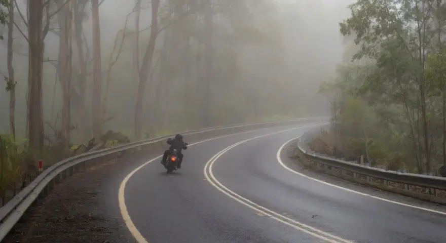 Motocyklista pokonuje zakręt na mokrej drodze, gdzie grozi mu aquaplaning. Mgła otula las, tworząc tajemniczą atmosferę.