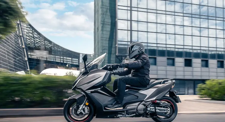 Mężczyzna w kasku jedzie na skuterze Kymco AK 550 obok nowoczesnego budynku.