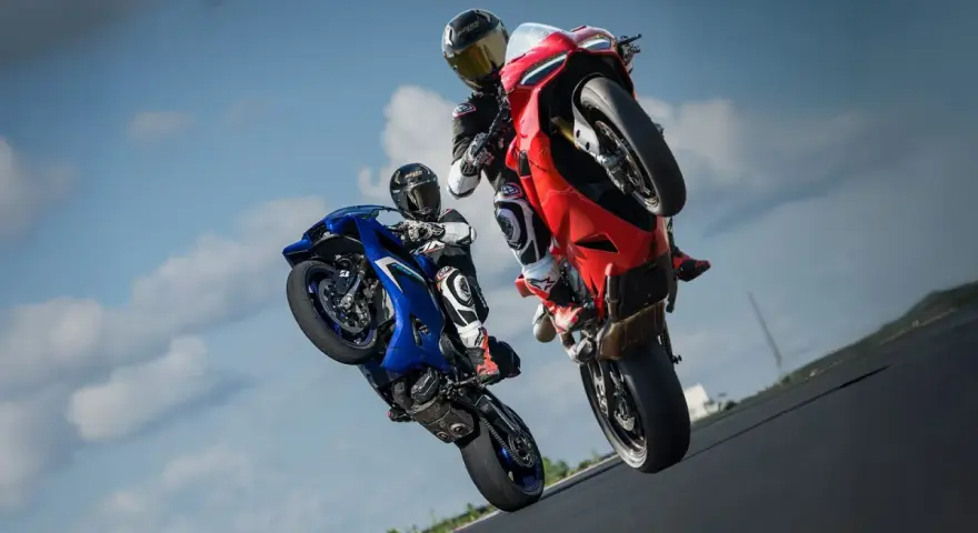 Dwóch motocyklistów na torze wyścigowym wykonuje wheelie. Jeden na niebieskim motocyklu, drugi na czerwonym Ducati Panigale V2.