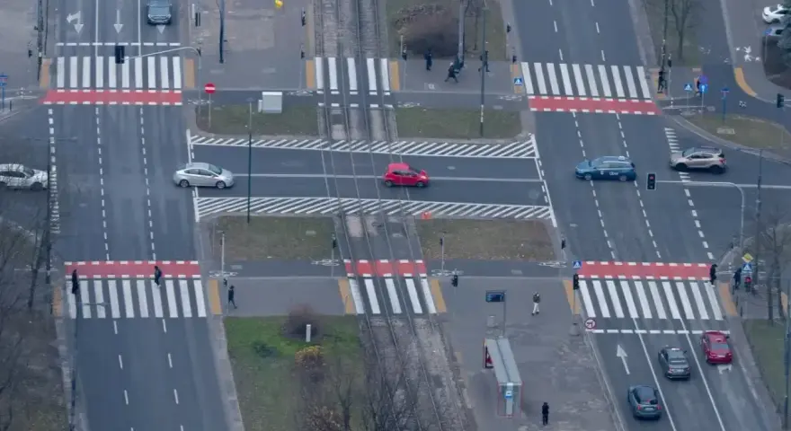 Skrzyżowanie z tramwajami i pasami. Czerwony samochód wykonuje wyprzedzanie na pasach, ryzykując kolizję z pieszymi.