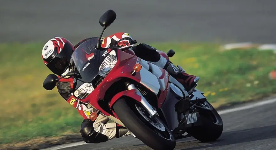 Motocyklista na torze pochyla się na swoim czerwono-białym motocyklu Yamaha R6 2001, w pełni ubrany w kombinezon i kask.