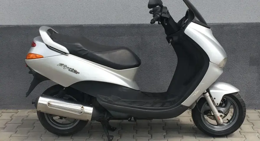 Srebrny skuter Peugeot Elyseo 125 z czarnym siedzeniem i przyciemnianą szybą, zaparkowany na chodniku.