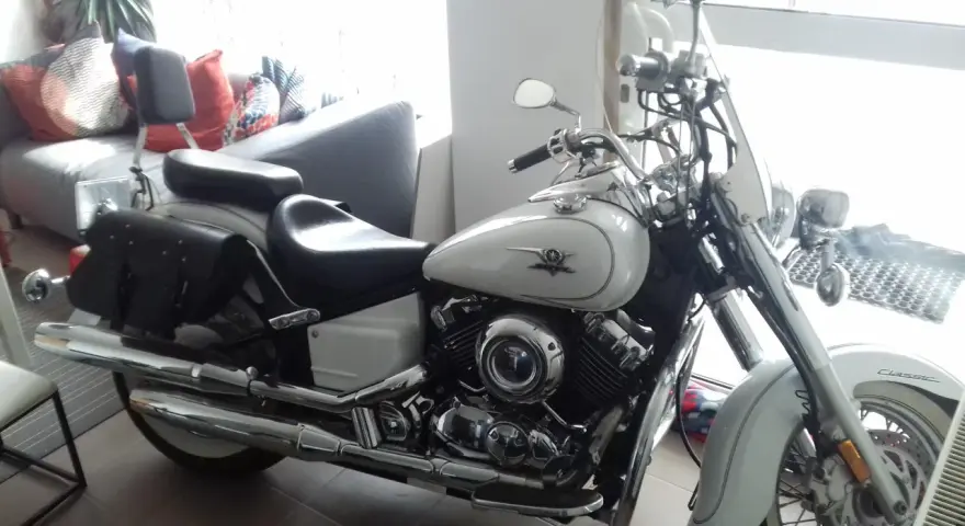 Biały motocykl Yamaha Drag Star 650, z sakwami i szybą. Wady i zalety tego klasyka są dyskutowane w tle, gdzie widać kanapę.