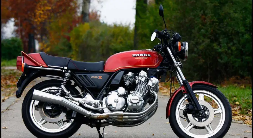 Klasyczny motocykl Honda CBX 1000 w kolorze czerwonym, z widocznym potężnym silnikiem i chromowanymi elementami, zaparkowany na tle jesiennej zieleni.