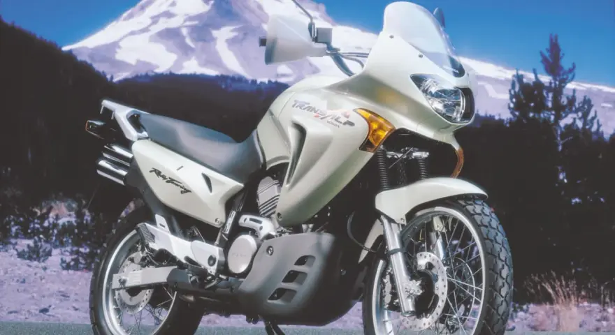 Srebrny motocykl Honda Transalp 650 gotowy na przygodę na tle ośnieżonej góry i lasu.