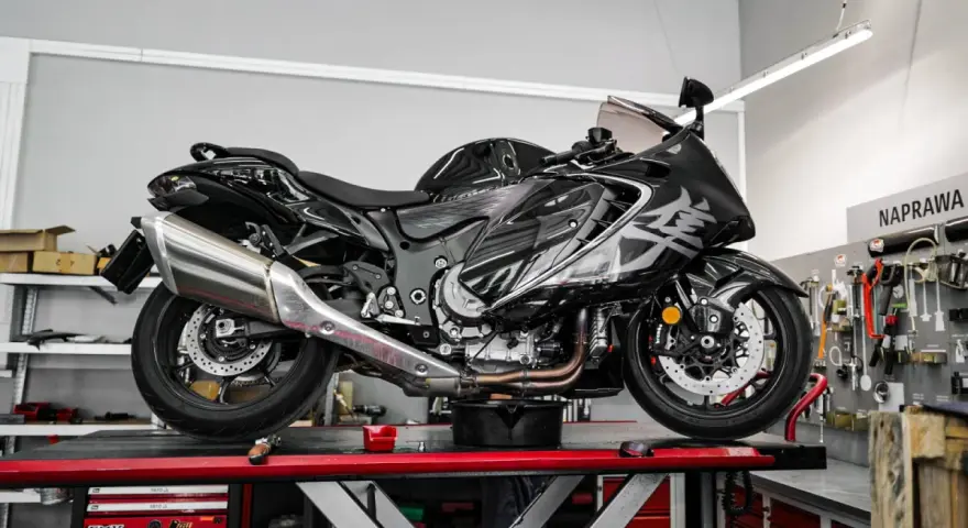 Czarny, lśniący motocykl typu naked bike na podnośniku w warsztacie. W tle tablica z narzędziami i napis "NAPRAWA".