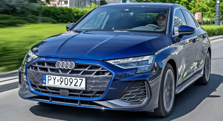 Młody kierowca w czapce za kierownicą granatowego Audi A3, jadącego po miejskiej drodze.