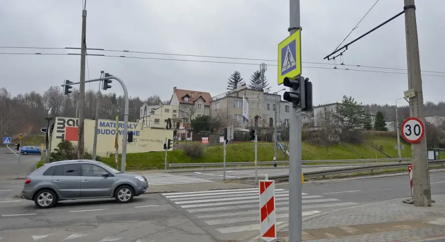 Szare auto zbliża się do przejścia dla pieszych, gdzie światła kolizyjne regulują ruch. W tle budynki i drzewa.