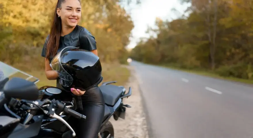Uśmiechnięta kobieta w skórzanym stroju trzyma kask obok swojego motocykla. Idealne motory dla kobiet na jesienną przejażdżkę.