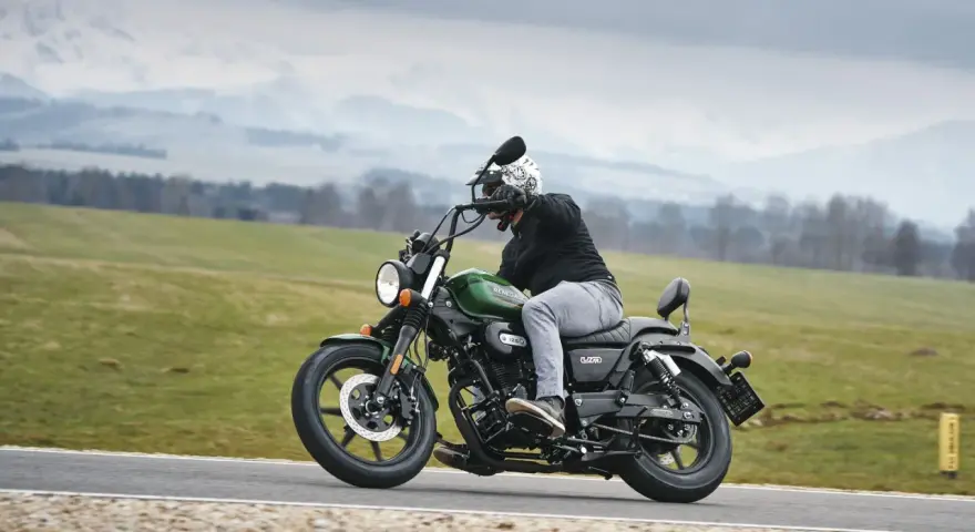 Motocyklista na zielonym chopperze UM Renegade 125 przemierza malowniczą drogę wśród zielonych pól i gór.