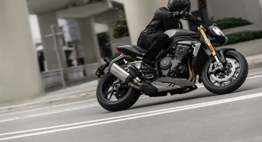 Motocyklista w kasku i skórzanej kurtce pochyla się na swoim motocyklu Triumph Speed Triple, pokonując zakręt na miejskiej drodze.