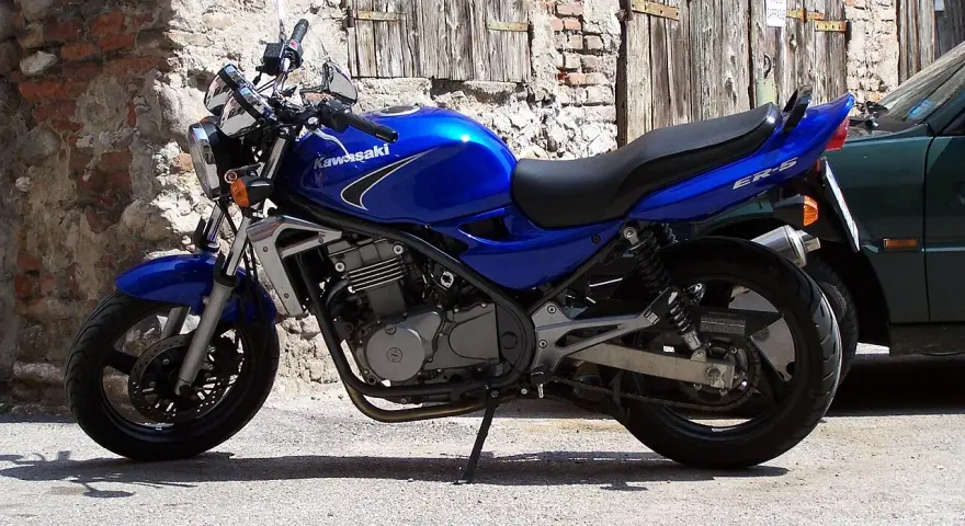 Błękitny motocykl Kawasaki ER-5 zaparkowany przy kamiennym murze i drewnianej bramie.