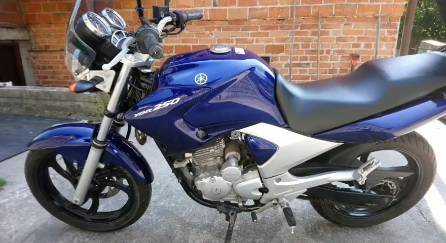 Błękitny motocykl Yamaha YBR 250 z chromowanymi elementami, zaparkowany na tle ceglanej ściany.