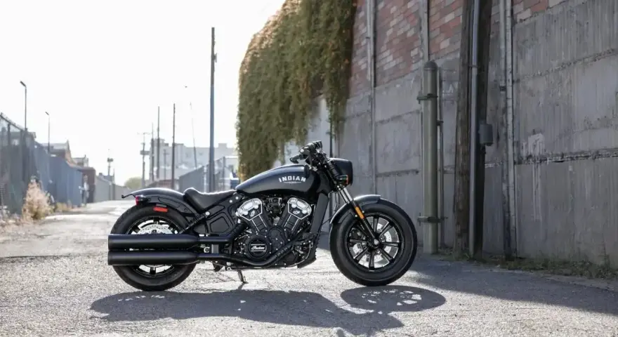 Czarny motocykl typu cruiser Indian Scout stoi na ulicy, z zielonym bluszczem zwisającym ze ściany w tle.