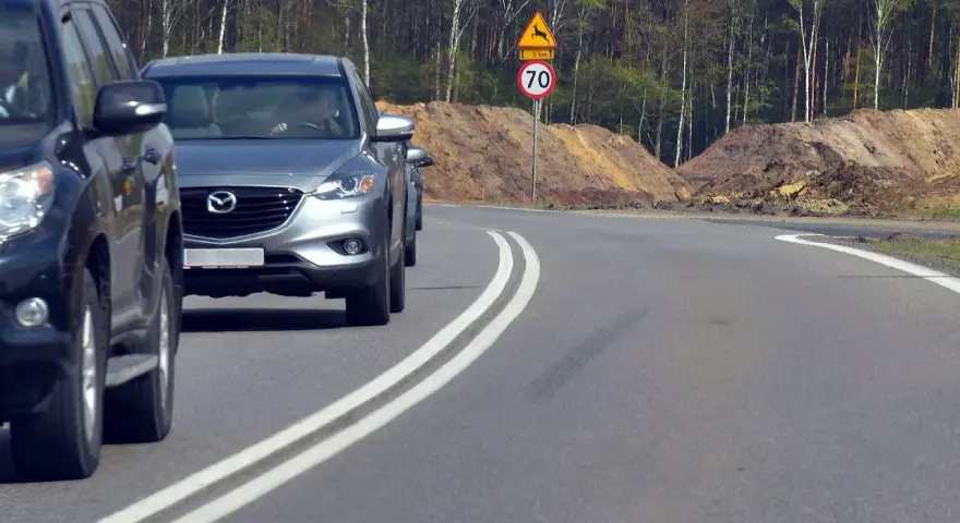 Kierowca próbuje wyprzedzanie na podwójnej ciągłej, ignorując znak ograniczenia prędkości do 70 km/h i ostrzeżenie o dzikich zwierzętach.