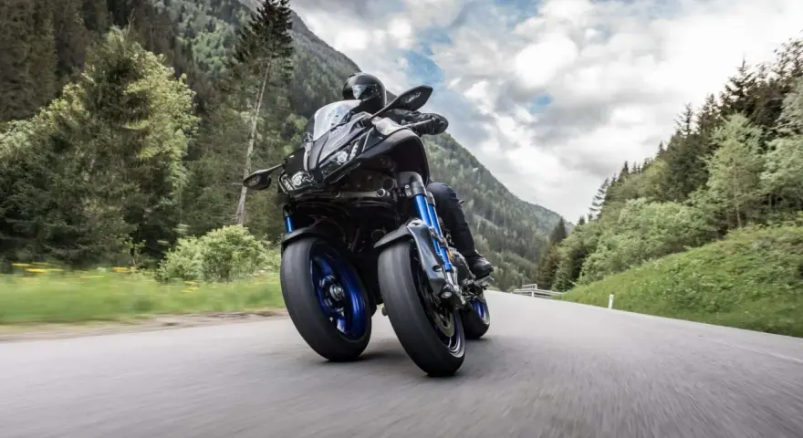 Motocyklista na Yamaha Niken pokonuje zakręt na górskiej drodze, otoczony zielonymi lasami i skalistymi szczytami.
