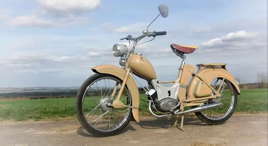 Beżowy motorower z NRD, z charakterystycznym siodełkiem i błotnikami, stoi na tle zielonych pól i błękitnego nieba.