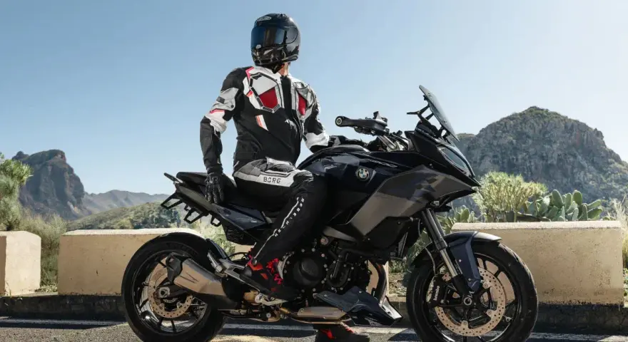 Motocyklista w kombinezonie na czarnym motocyklu BMW, gotowy do podróży po górskich drogach. Wśród wielu marek motocykli, ta prezentuje się imponująco.