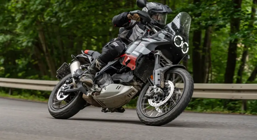 Motocyklista w kasku i stroju terenowym pochyla się na zakręcie na drodze leśnej, prowadząc czarny motocykl Ducati DesertX.